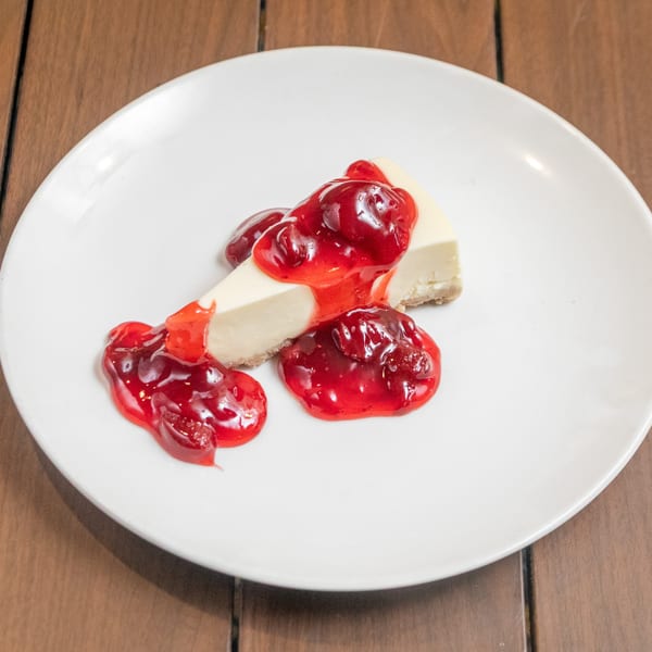 A slice of cheesecake with a graham cracker crust, topped with bright red cherry sauce, on a white plate.