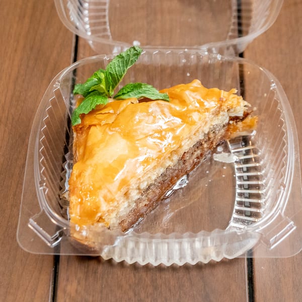 A slice of baklava with a mint garnish in a clear plastic container on a wooden table.